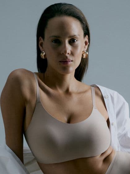 A woman poses confidently in a beige bra and open white shirt. She is wearing large gold earrings and looking directly at the camera.