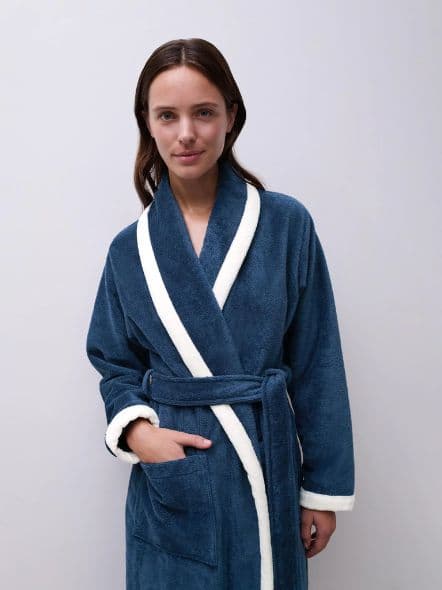 Woman wearing a blue bathrobe with white trim, standing against a plain background.