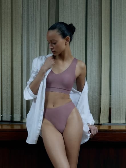 A woman in rose colored lingerie and an open white shirt stands by a wood-paneled wall with vertical blinds, looking down.